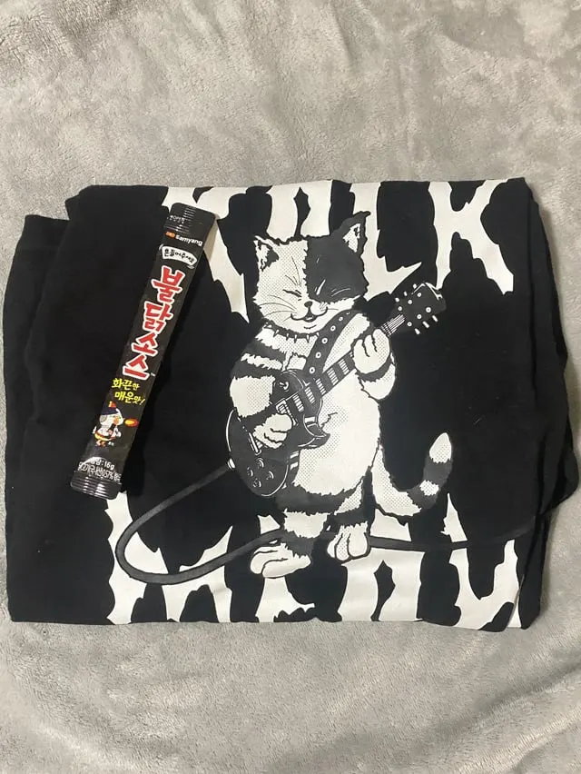 Black t-shirt printed with a cat playing an electric guitar, beside a Korean snack stick